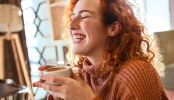 Cozy woman with curly red hair enjoying a warm coffee, smiling happily indoors with sunlight streaming through the window.