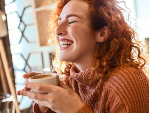 Cozy woman with curly red hair enjoying a warm coffee, smiling happily indoors with sunlight streaming through the window.