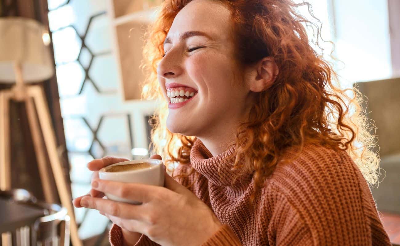 Cozy woman with curly red hair enjoying a warm coffee, smiling happily indoors with sunlight streaming through the window.