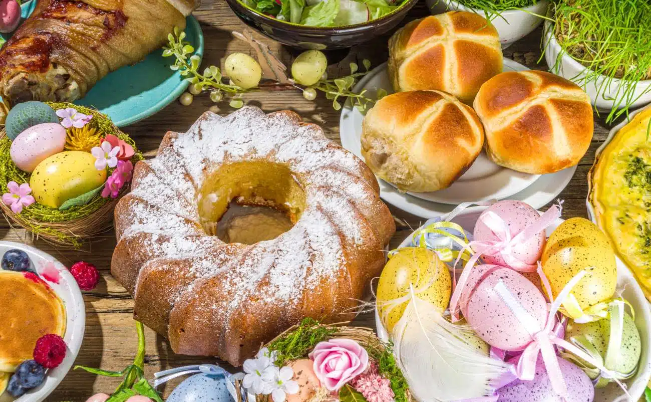 Easter-themed food spread with decorated eggs, baked bread rolls, a cake dusted with powdered sugar, fresh salad, and various colorful dishes on a rustic wooden table.