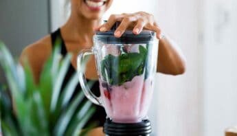 Blender containing green leafy vegetables and pink smoothie, with smiling woman in background, promoting healthy living and nutritious smoothies for better health.