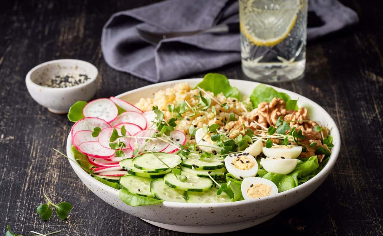 Fresh vegetable salad with radishes, cucumbers, boiled eggs, walnuts, and microgreens, served on a white bowl with a glass of water and seasoning on a rustic dark wooden table.