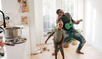 Father playing guitar and singing with daughter dancing in a bright home, creating a joyful and healthy family activity.