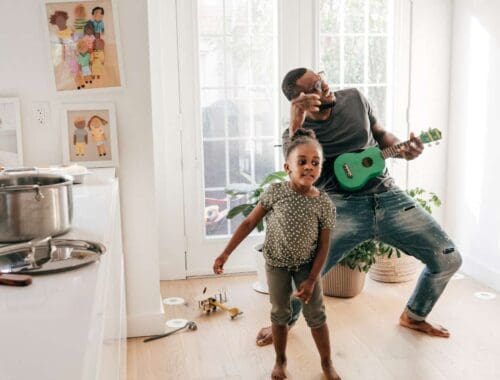 Father playing guitar and singing with daughter dancing in a bright home, creating a joyful and healthy family activity.