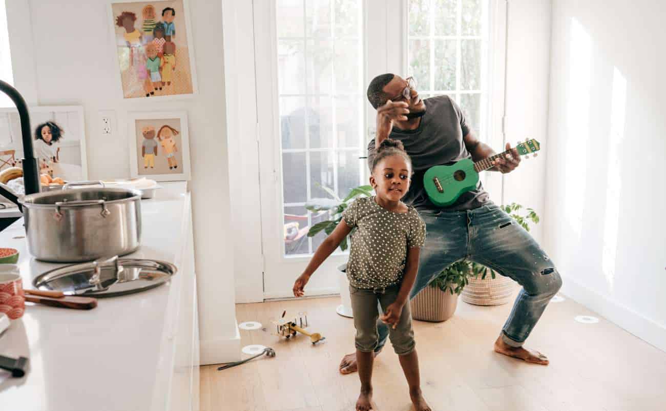 Father playing guitar and singing with daughter dancing in a bright home, creating a joyful and healthy family activity.