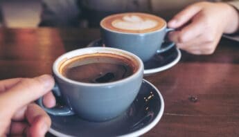Rich, steaming cups of coffee served in gray ceramic cups on a wooden table, with one person holding a cup and another reaching for their coffee, suggesting a relaxing coffee break or social moment.