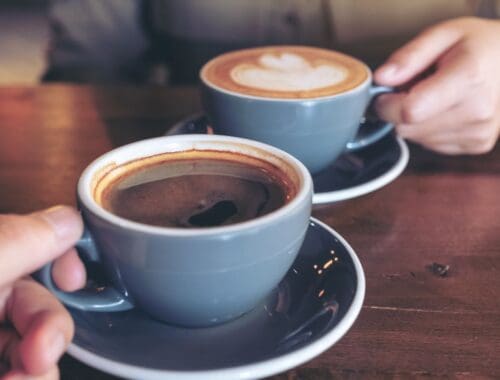 Rich, steaming cups of coffee served in gray ceramic cups on a wooden table, with one person holding a cup and another reaching for their coffee, suggesting a relaxing coffee break or social moment.