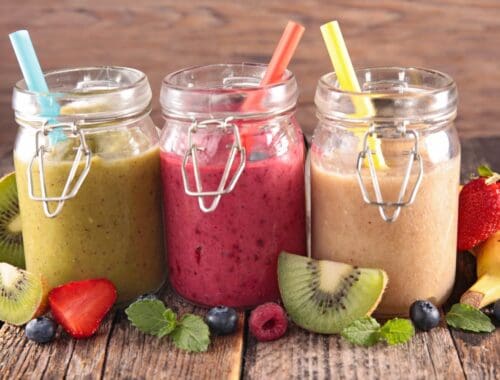 Refreshing fruit smoothies in glass jars with colorful straws on a rustic wooden surface, surrounded by fresh fruits like strawberries, kiwi, blueberries, raspberries, and bananas. Healthy and delicious beverage options for a nutritious lifestyle.