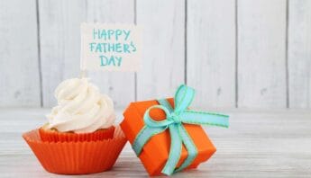Delicious cupcake with white frosting and a "Happy Father's Day" note on top, placed next to an orange gift box with a green ribbon, celebrating Father's Day with sweet treats and thoughtful presents.