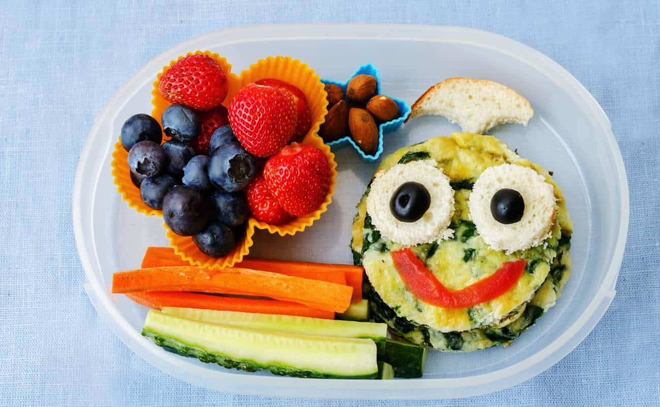 Fresh fruit and vegetable snack plate with blueberries, strawberries, carrots, cucumbers, nuts, and a smiling face-shaped egg dish with olives and red bell pepper for a healthy meal.