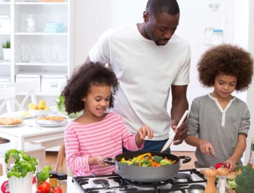 Fresh vegetables cooking in a skillet with a dad and two daughters in a bright, modern kitchen promoting healthy family eating and nutritious meal preparation.