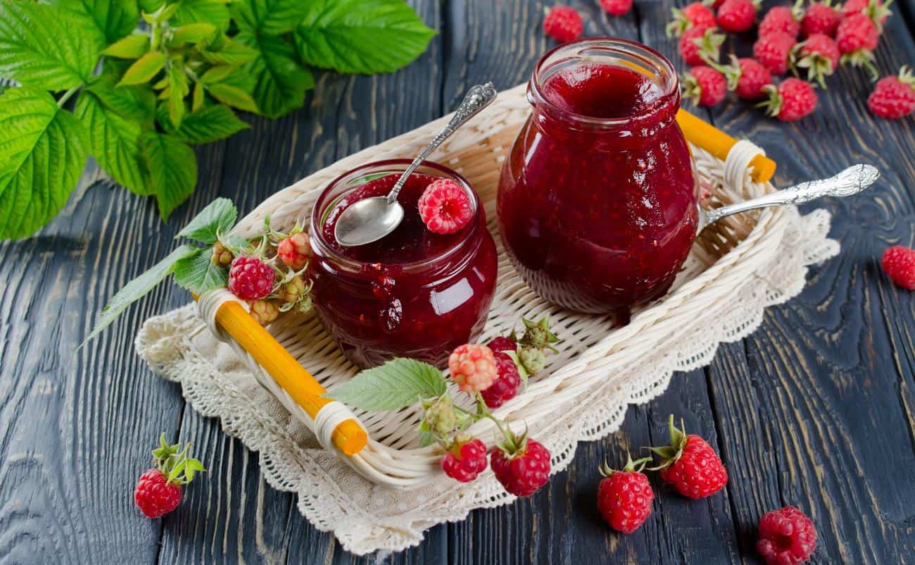 Fresh homemade raspberry jam in glass jars with raspberries and green leaves on a rustic wooden table for healthy eating and natural preserves.