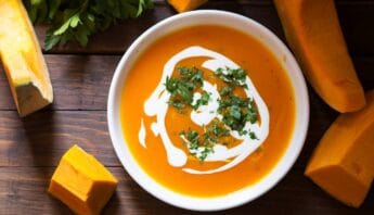 Creamy pumpkin soup garnished with cilantro and a swirl of sour cream, surrounded by fresh pumpkin pieces on a rustic wooden surface. Perfect for a healthy, nutritious meal.
