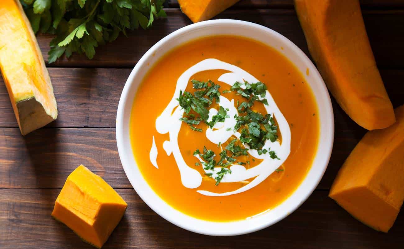 Creamy pumpkin soup garnished with cilantro and a swirl of sour cream, surrounded by fresh pumpkin pieces on a rustic wooden surface. Perfect for a healthy, nutritious meal.