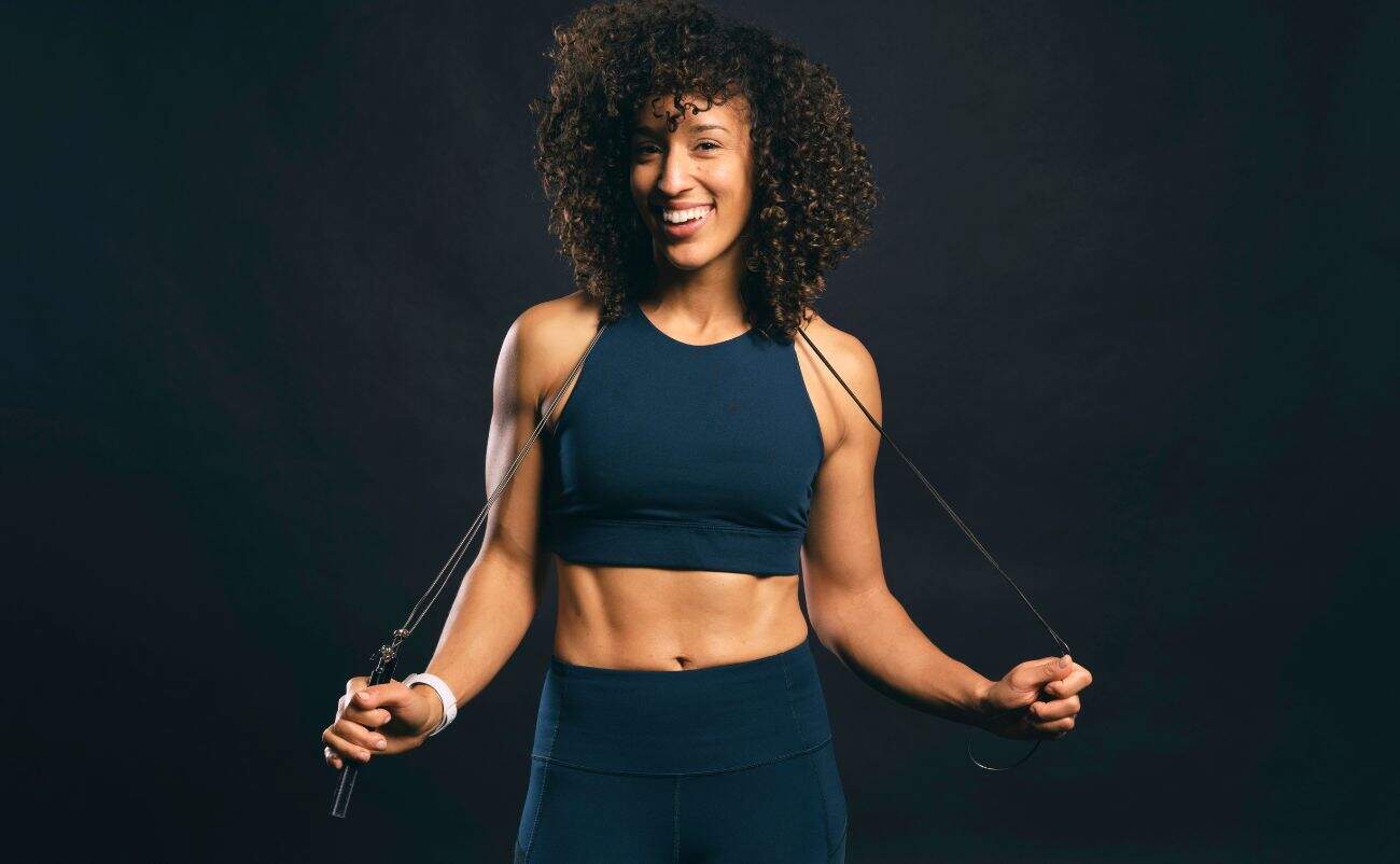 Fit woman smiling holding skipping rope, athletic wear, fitness, health, workout, exercise, active lifestyle, black background, motivational sportswear.