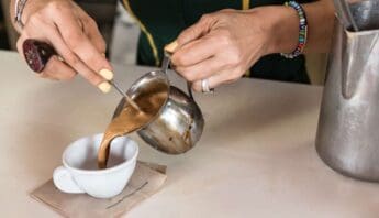 Hot chocolate being poured into a small white cup from a metal pitcher, with a person wearing colorful beaded bracelets and a large ring on their hand.