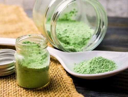 Powdered green superfood supplement in glass jars with a white spoon, on rustic burlap and wooden table, promoting health and fitness.