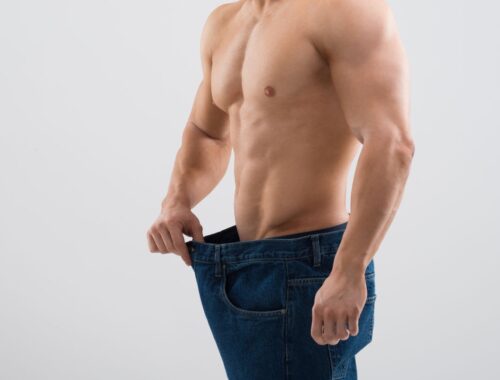 Slim fit man holding out oversized jeans waistband, emphasizing weight loss and body transformation.