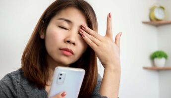 Worried woman holding a smartphone, touching her forehead, possibly experiencing stress or headache, indoors with a minimal background.