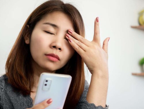Worried woman holding a smartphone, touching her forehead, possibly experiencing stress or headache, indoors with a minimal background.