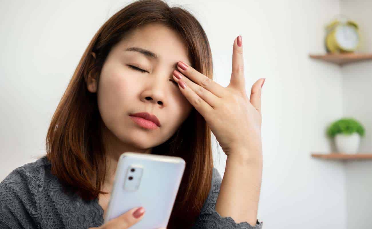 Worried woman holding a smartphone, touching her forehead, possibly experiencing stress or headache, indoors with a minimal background.