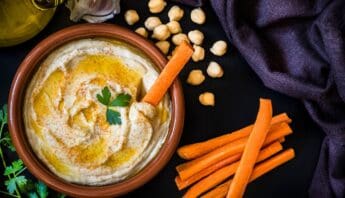 Mediterranean Diet Dessert Recipes for Weight Loss 7 Creamy hummus in a terracotta bowl garnished with parsley, surrounded by carrot sticks, chickpeas, garlic, olive oil, and fresh herbs on a dark surface.