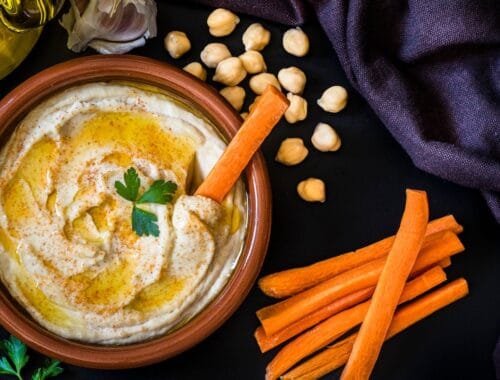 Creamy hummus in a terracotta bowl garnished with parsley, surrounded by carrot sticks, chickpeas, garlic, olive oil, and fresh herbs on a dark surface.