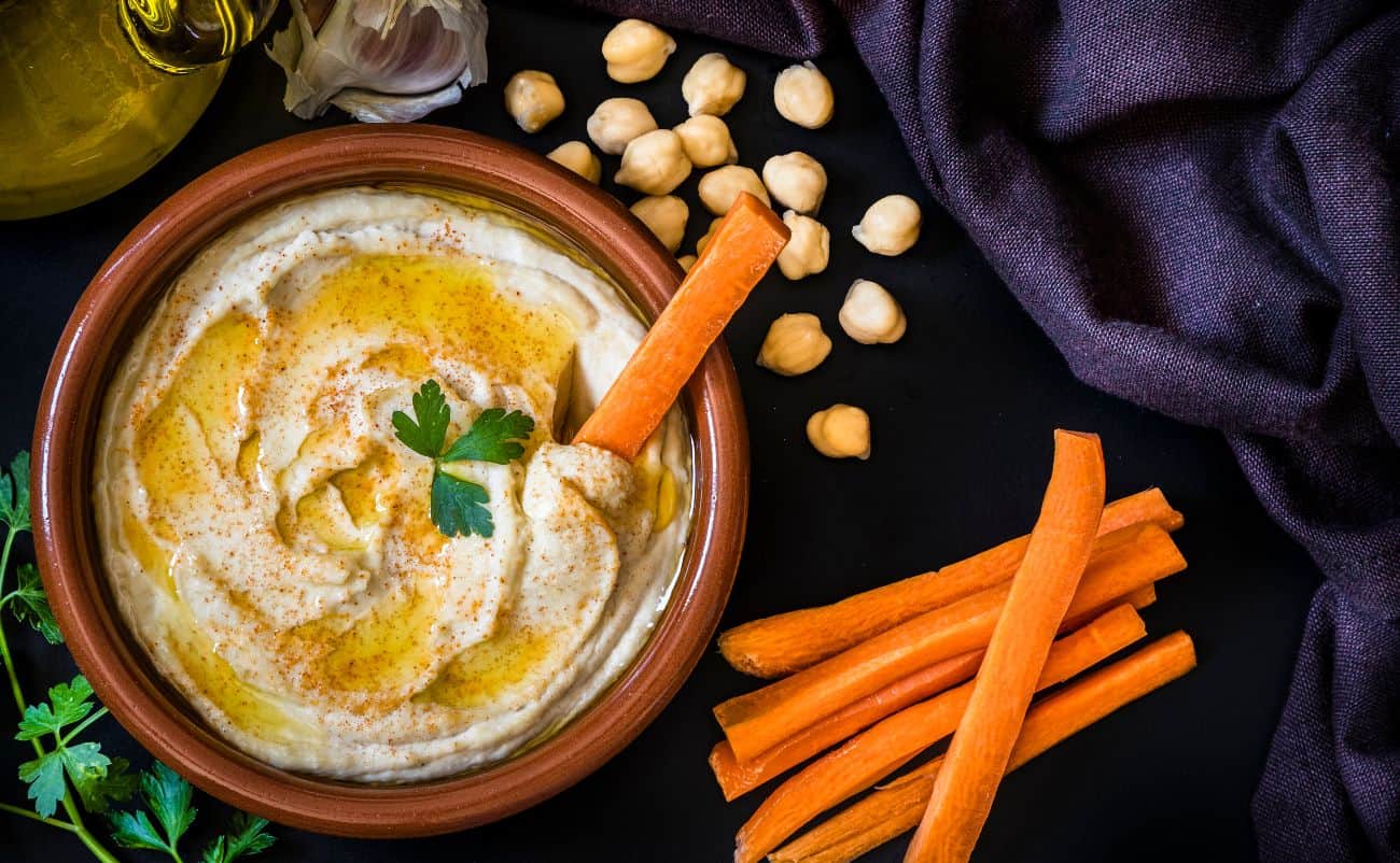 Creamy hummus in a terracotta bowl garnished with parsley, surrounded by carrot sticks, chickpeas, garlic, olive oil, and fresh herbs on a dark surface.