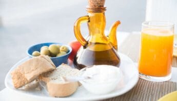 Organic olive oil in a glass bottle with a cork stopper, served alongside bread, olives, and orange juice on a breakfast table.