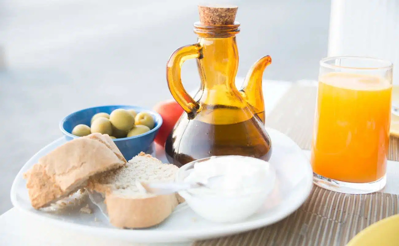 Kickstart Your Health: The Ultimate 14-Day Mediterranean Diet Meal Plan for Beginners! 1 Organic olive oil in a glass bottle with a cork stopper, served alongside bread, olives, and orange juice on a breakfast table.