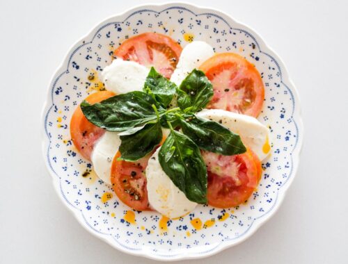Fresh Caprese salad with ripe tomatoes, mozzarella, basil leaves, olive oil, and black pepper on a decorative white plate.