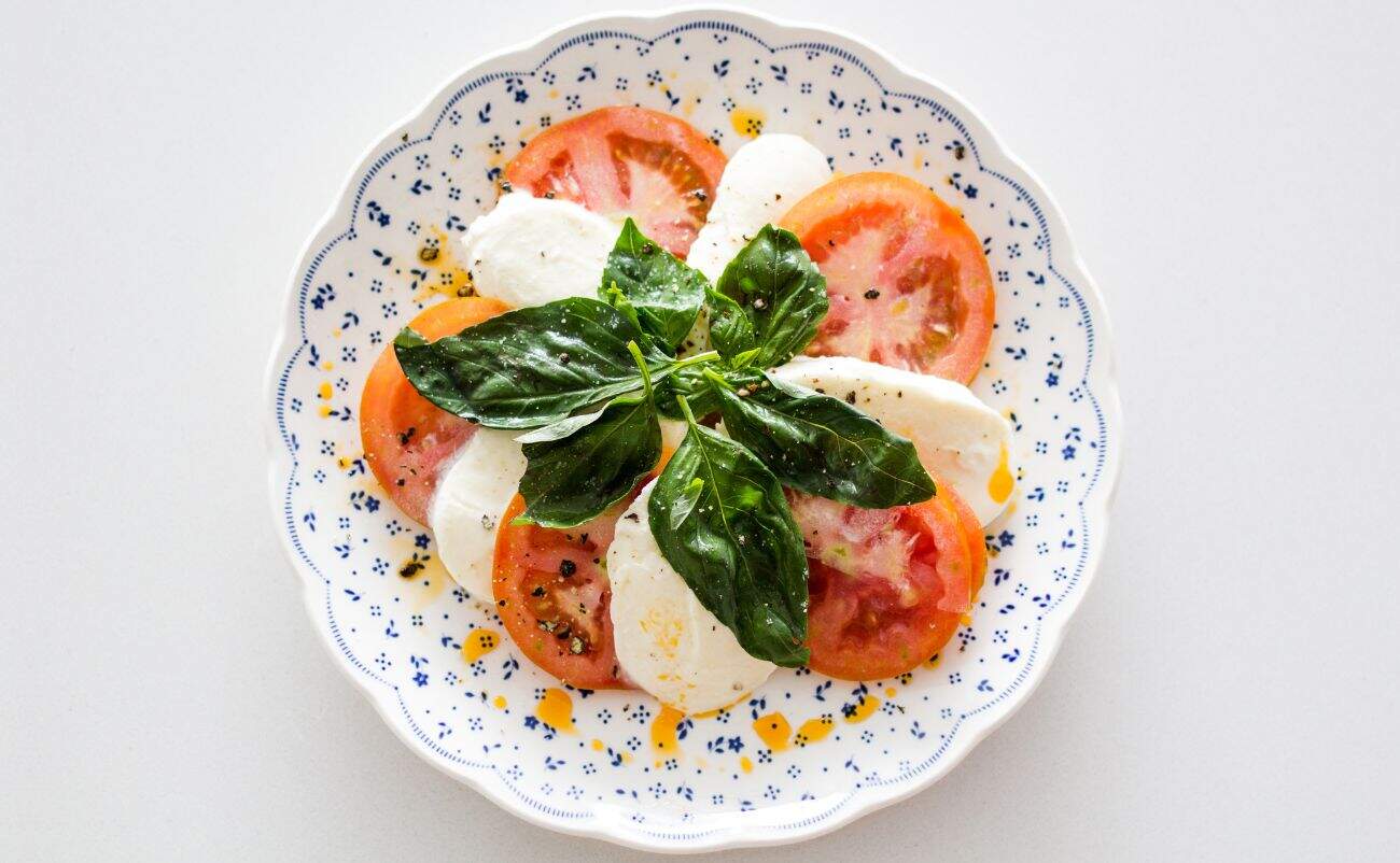 Fresh Caprese salad with ripe tomatoes, mozzarella, basil leaves, olive oil, and black pepper on a decorative white plate.