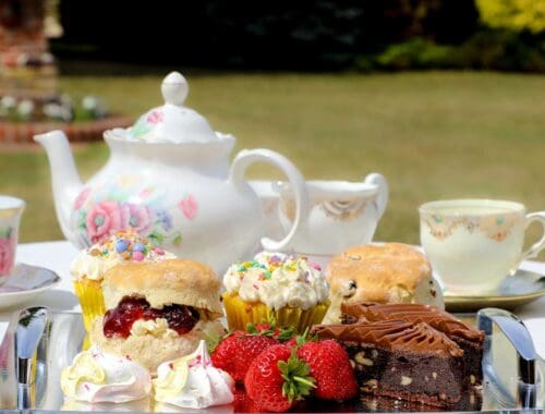 Tea party with assorted desserts, cupcakes, strawberries, and fine china on a outdoor table in a garden setting.
