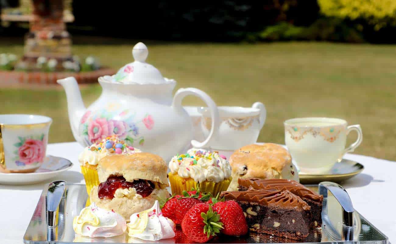 Tea party with assorted desserts, cupcakes, strawberries, and fine china on a outdoor table in a garden setting.
