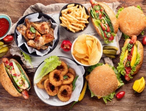 Rich, flavorful fast food meal with burgers, fries, fried onions, and chicken wings on a rustic wooden table with vegetables and dipping sauces.