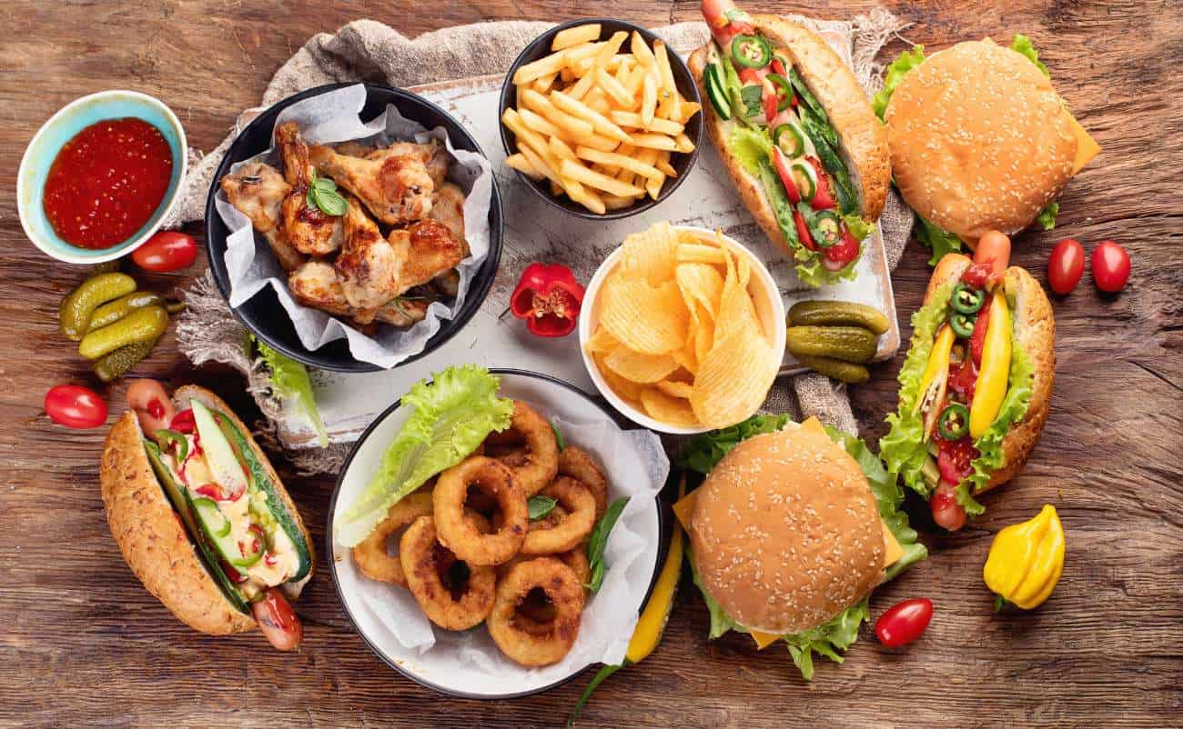 Rich, flavorful fast food meal with burgers, fries, fried onions, and chicken wings on a rustic wooden table with vegetables and dipping sauces.