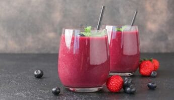 Refreshing berry smoothies topped with fresh mint in clear glasses with black straws, surrounded by strawberries and blueberries on a dark surface.