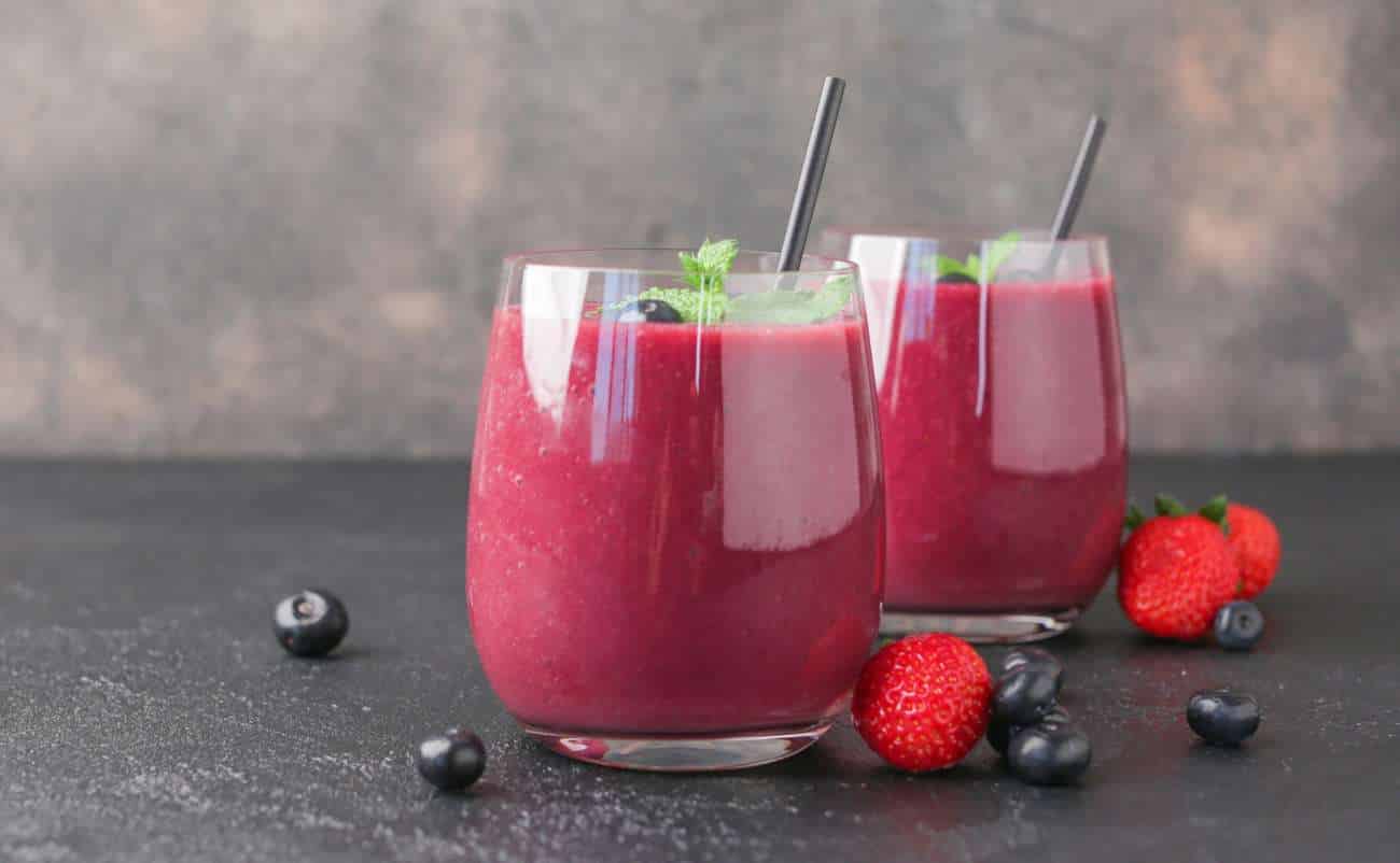 Refreshing berry smoothies topped with fresh mint in clear glasses with black straws, surrounded by strawberries and blueberries on a dark surface.