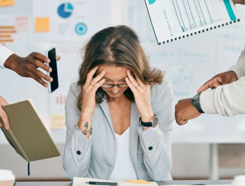 Overwhelmed woman at work stressed with multiple hands reaching out holding phones and documents, illustrating burnout or work-related stress.