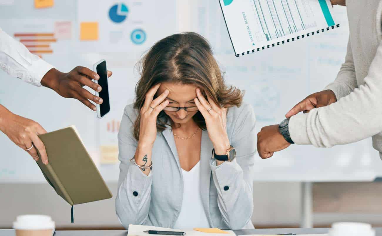 Overwhelmed woman at work stressed with multiple hands reaching out holding phones and documents, illustrating burnout or work-related stress.