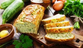 Savoury zucchini bread loaf with sliced pieces on rustic wooden cutting board, surrounded by fresh vegetables, eggs, and herbs, emphasizing healthy homemade food options for balanced nutrition.