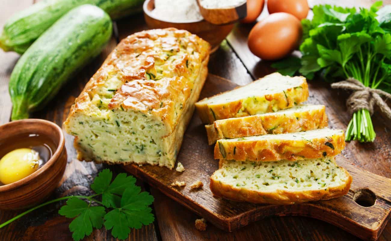 Savoury zucchini bread loaf with sliced pieces on rustic wooden cutting board, surrounded by fresh vegetables, eggs, and herbs, emphasizing healthy homemade food options for balanced nutrition.