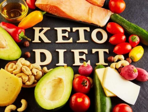 Keto diet foods including avocados, cherry tomatoes, cheese, cucumbers, and nuts arranged around the words "KETO DIET" on a dark surface. Healthy fats and vegetables promote low-carb, high-fat keto eating.