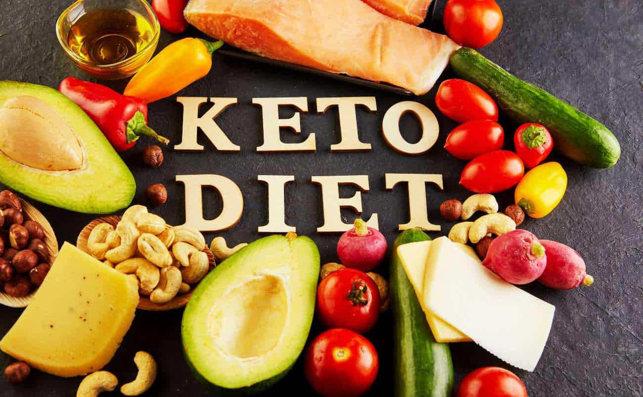 Keto diet foods including avocados, cherry tomatoes, cheese, cucumbers, and nuts arranged around the words "KETO DIET" on a dark surface. Healthy fats and vegetables promote low-carb, high-fat keto eating.