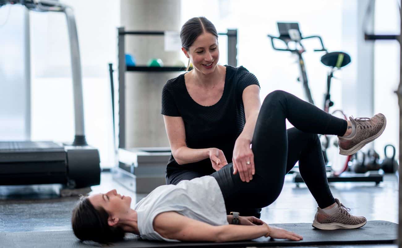 Women personal trainer helping woman with stretching exercise in gym.