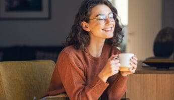 Relaxed woman enjoying coffee at home, smiling with eyes closed, cozy sweater, natural light, comfort, wellness, happiness, self-care, mental health, tranquility.