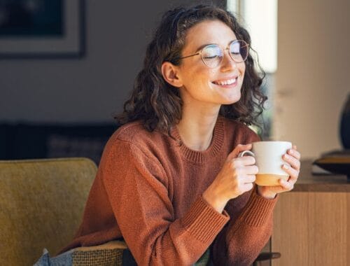 Relaxed woman enjoying coffee at home, smiling with eyes closed, cozy sweater, natural light, comfort, wellness, happiness, self-care, mental health, tranquility.