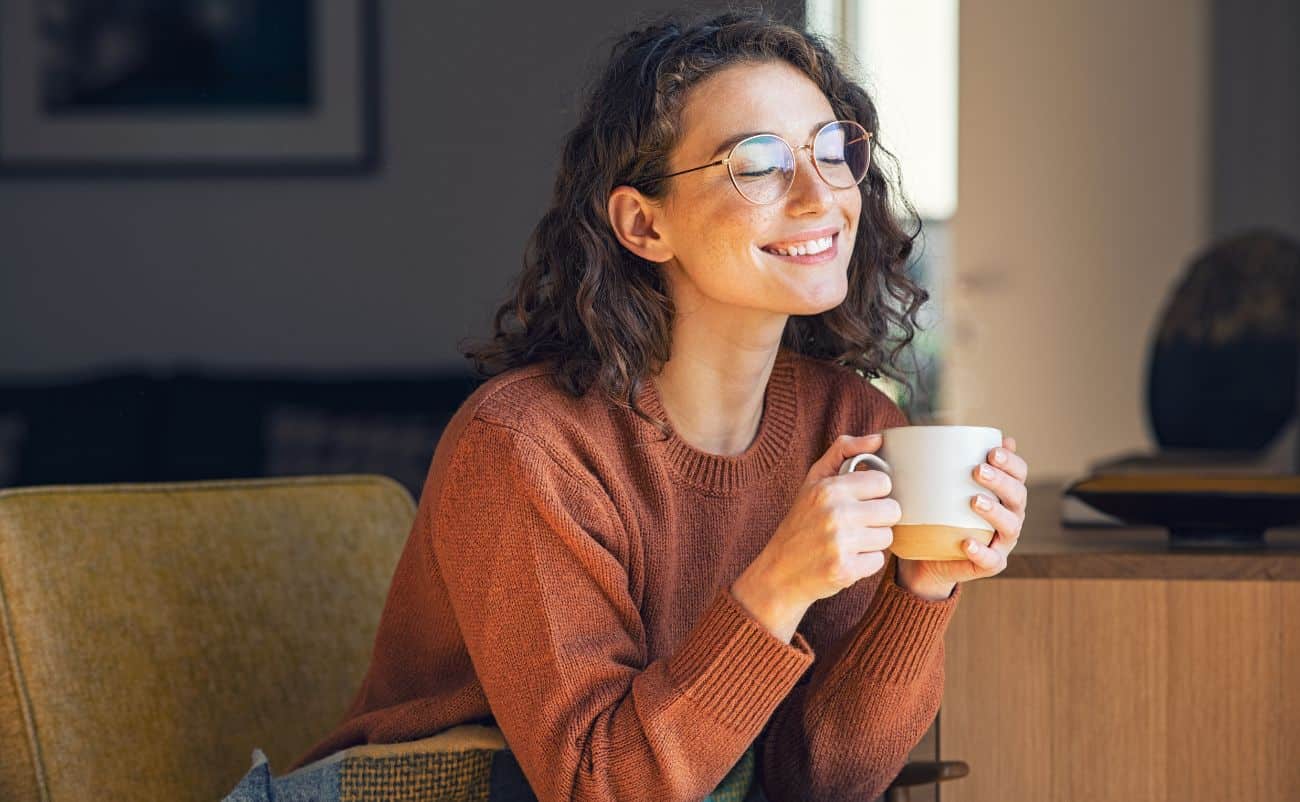 Relaxed woman enjoying coffee at home, smiling with eyes closed, cozy sweater, natural light, comfort, wellness, happiness, self-care, mental health, tranquility.