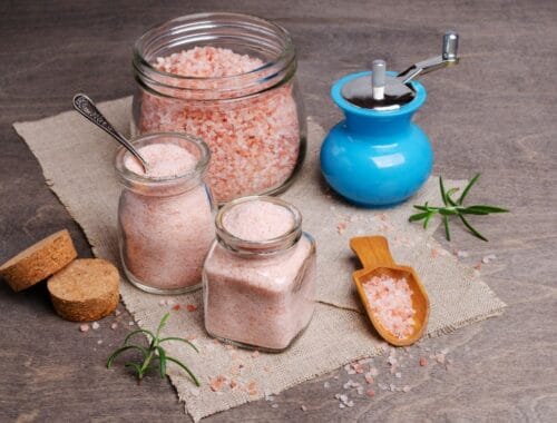 Pink Himalayan salt in jars and a salt grinder on a wooden surface for health and wellness.