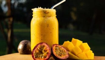 Fresh passion fruit and mango smoothie in a glass jar with a straw, displayed outdoors on a wooden surface.